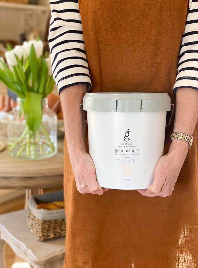 Bilden visar Grafenfärg Godnattpussfärg i kulören Morgondis i en vit burk med namnet på färgen på sidan. En kvinna med en randig t-shirt och en brun förkläde på sig håller i burken